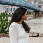 A woman in a white dress stands by the river with the John A. Roebling Suspension Bridge in the background.